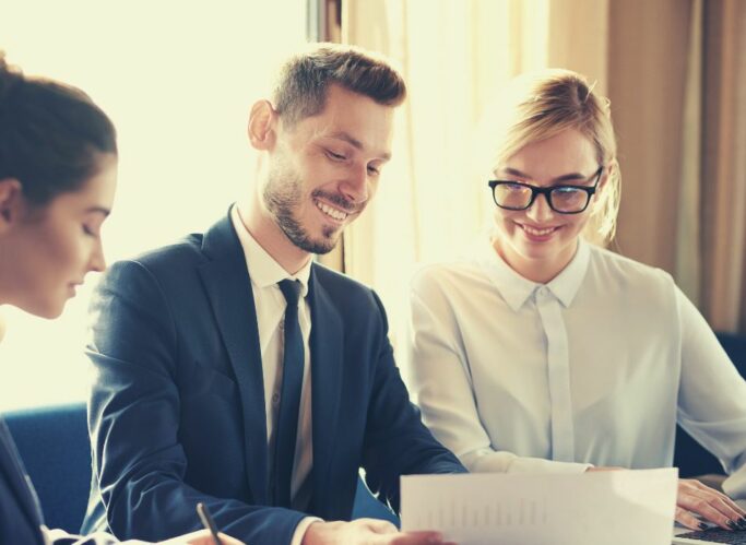Trois professionnels, deux femmes et un homme, sont assis à une table, examinant des documents en souriant. Vêtus de tenues professionnelles, ils semblent participer à une réunion collaborative avec leur courtier en assurance copropriété.