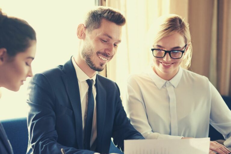 Trois professionnels, deux femmes et un homme, sont assis à une table, examinant des documents en souriant. Vêtus de tenues professionnelles, ils semblent participer à une réunion collaborative avec leur courtier en assurance copropriété.