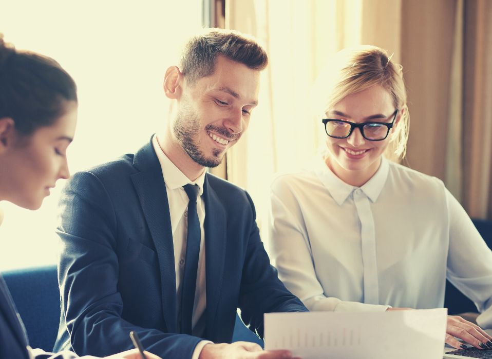 Trois professionnels, deux femmes et un homme, sont assis à une table, examinant des documents en souriant. Vêtus de tenues professionnelles, ils semblent participer à une réunion collaborative avec leur courtier en assurance copropriété.